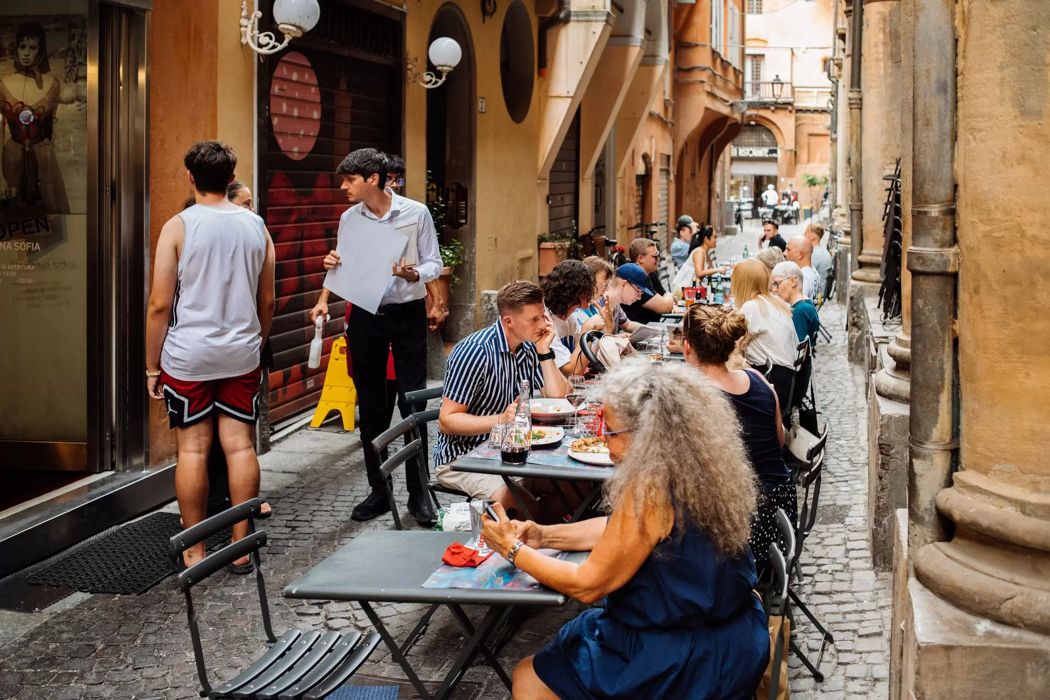 People sitting at tables on a narrow cobblestone street.