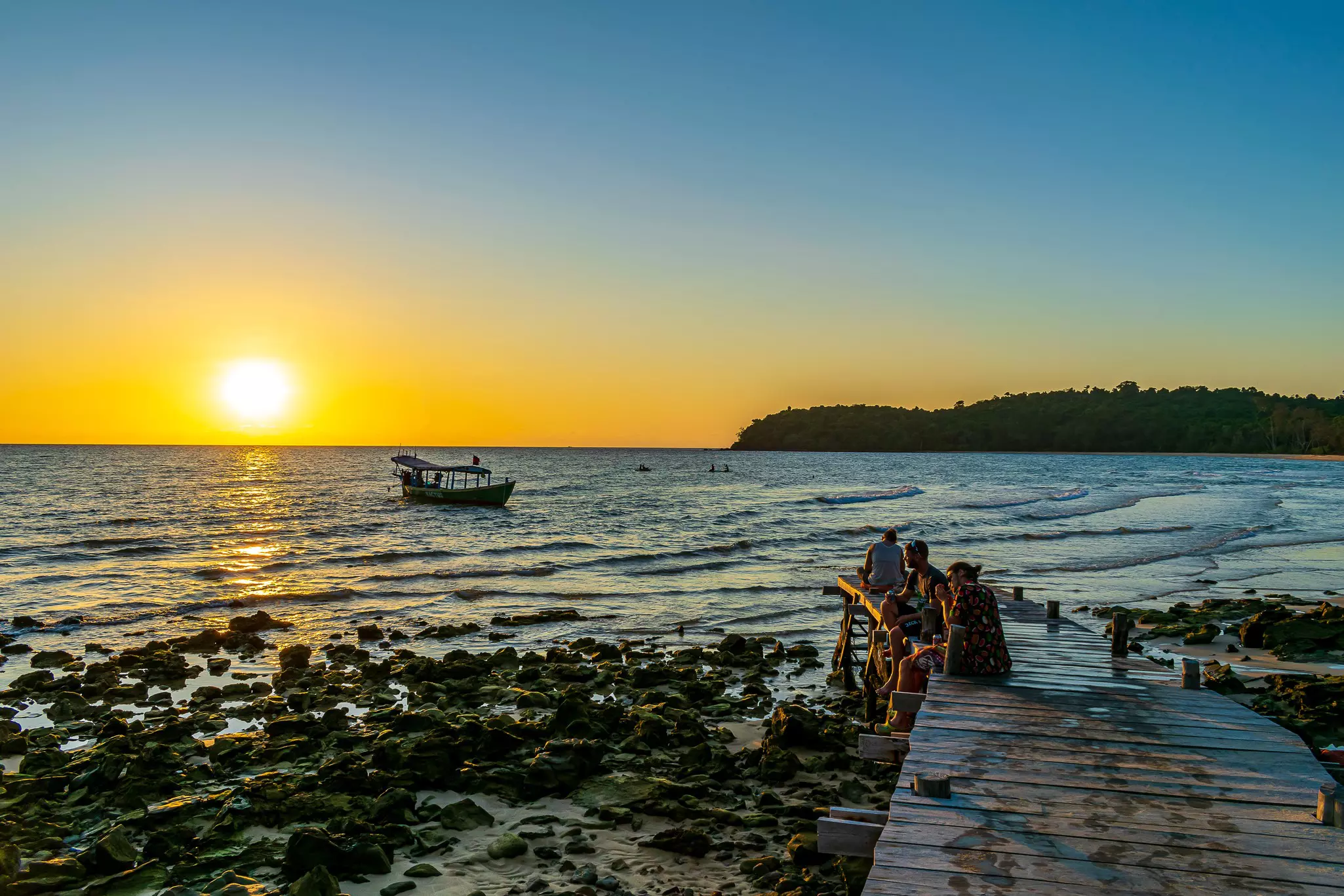 People sit on a short pier by a rocky shore looking at the sunset; there is a single boat in the water.