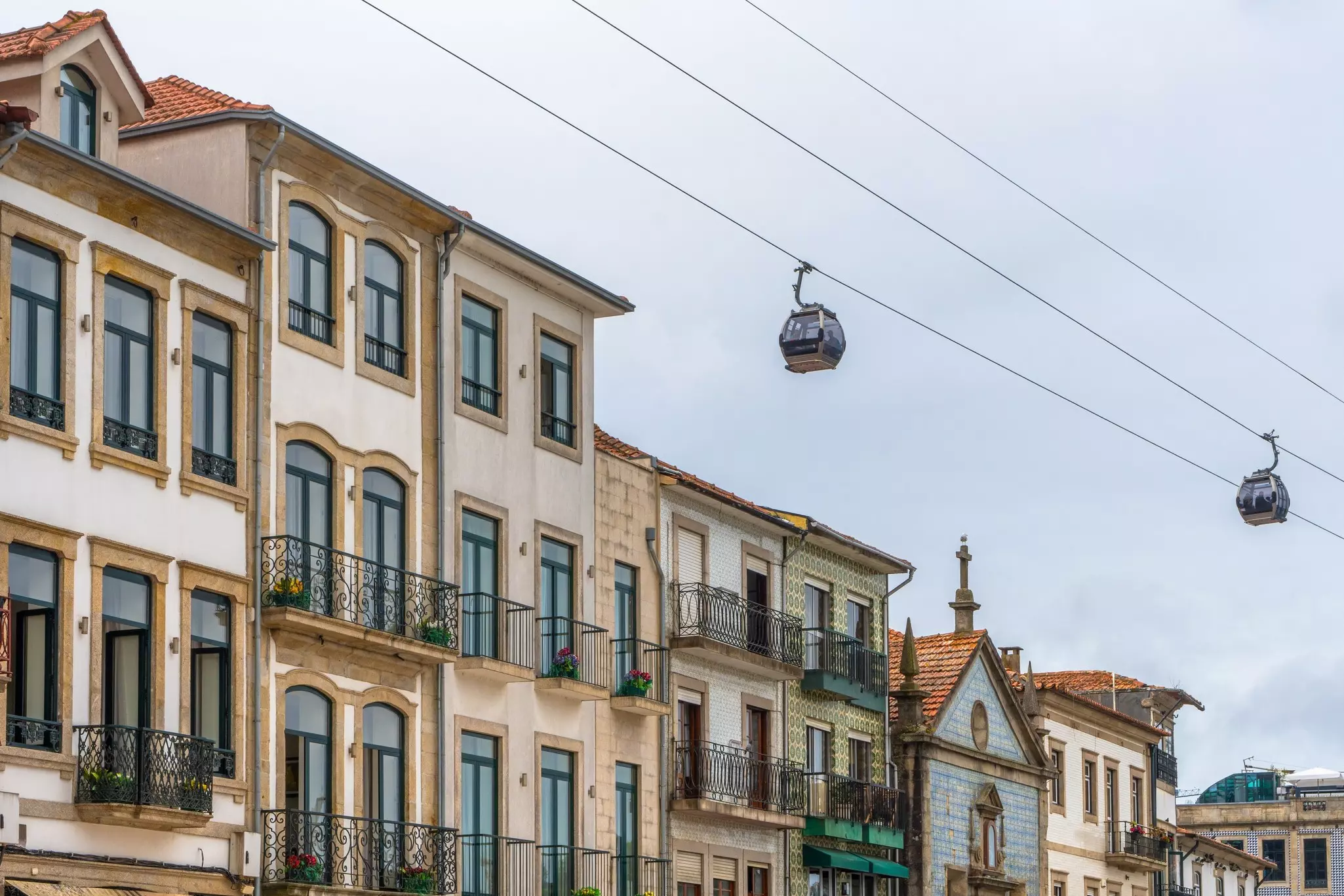 Two gondolas over buildings in Porto, Portugal, under a cloudy sky.