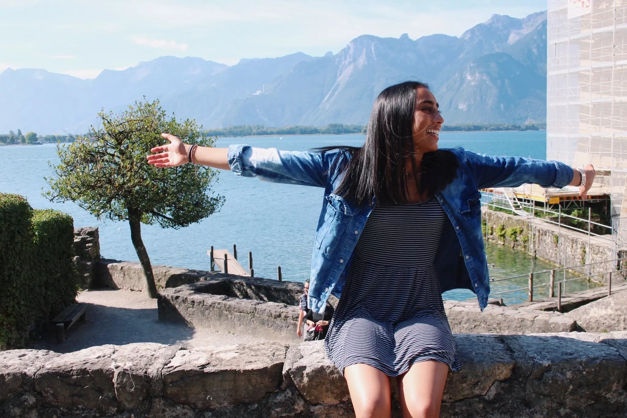 A woman sits with her arms outstretched beside a lake in Switzerland.