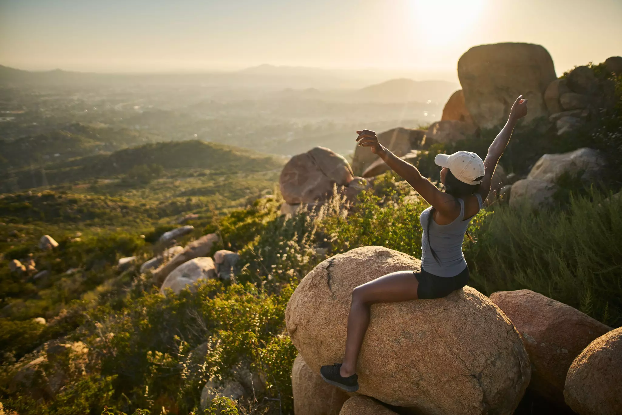 fit female hiker sitting on rock at mountain top looking at city in distance with arms up