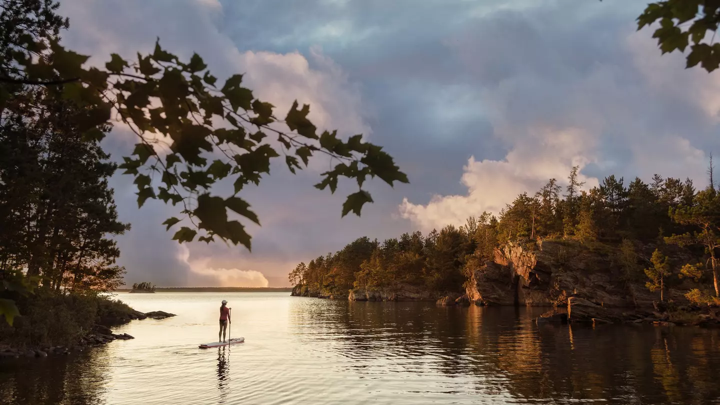 A solo figure on a paddleboard on a lake in a wooded area as the sky turns orange-pink at sunset