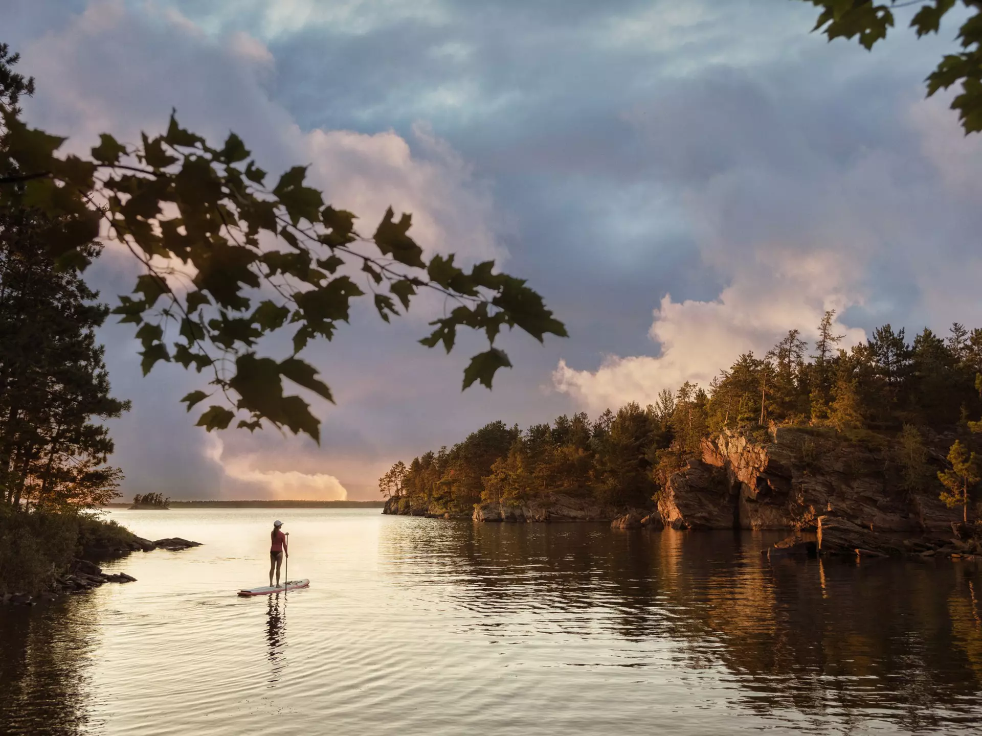 A solo figure on a paddleboard on a lake in a wooded area as the sky turns orange-pink at sunset