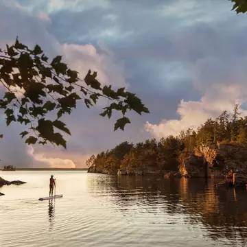 A solo figure on a paddleboard on a lake in a wooded area as the sky turns orange-pink at sunset
