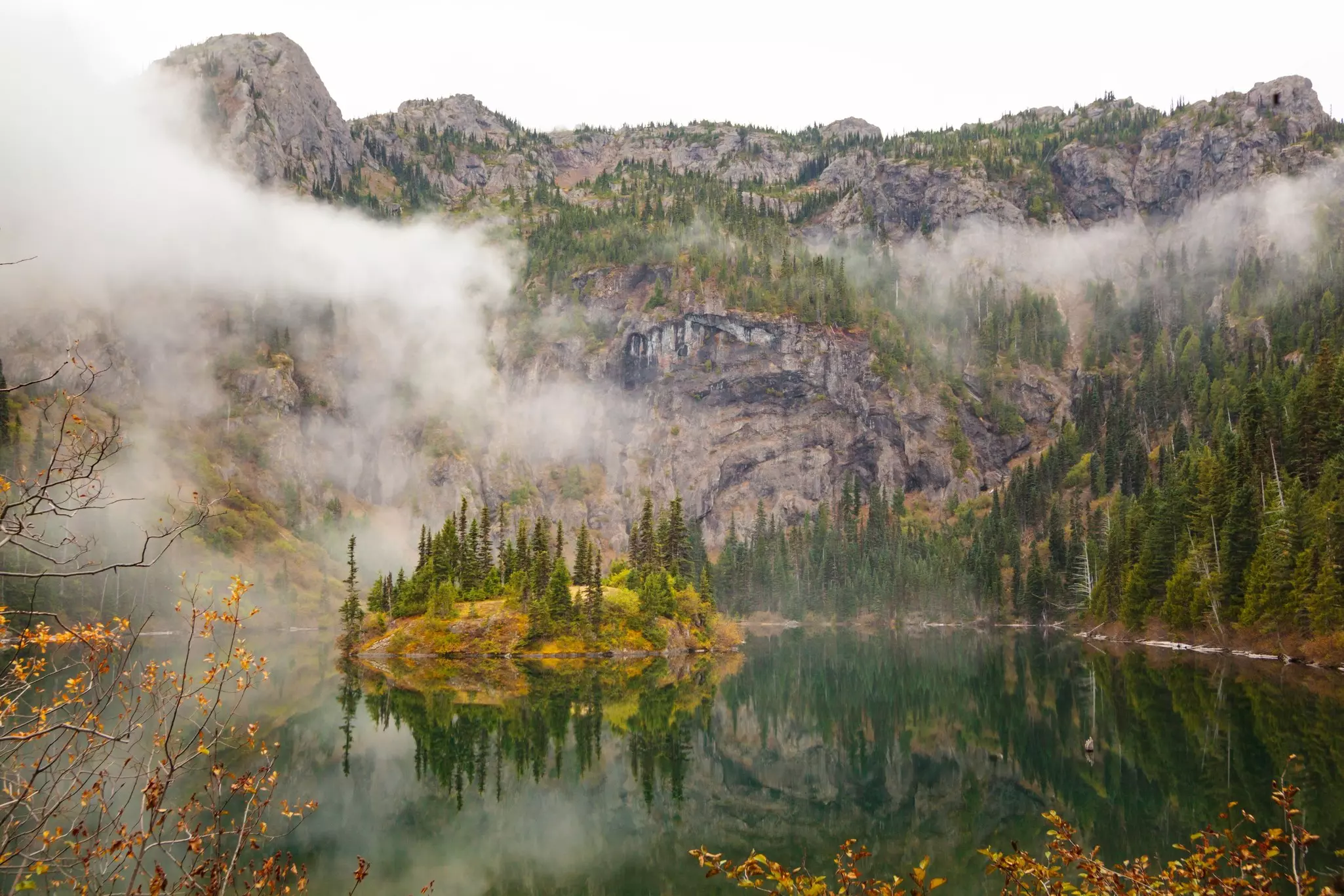 Rocky mountains enclose valley with a green lake; a low cloud is in the valley, and some trees are in autumn colors.