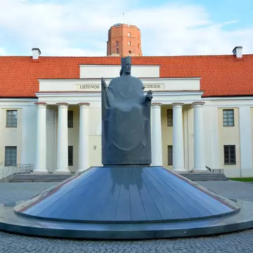 Seated dark granite king on a round plinth before a white columned building with orange roof, a brick tower beyond