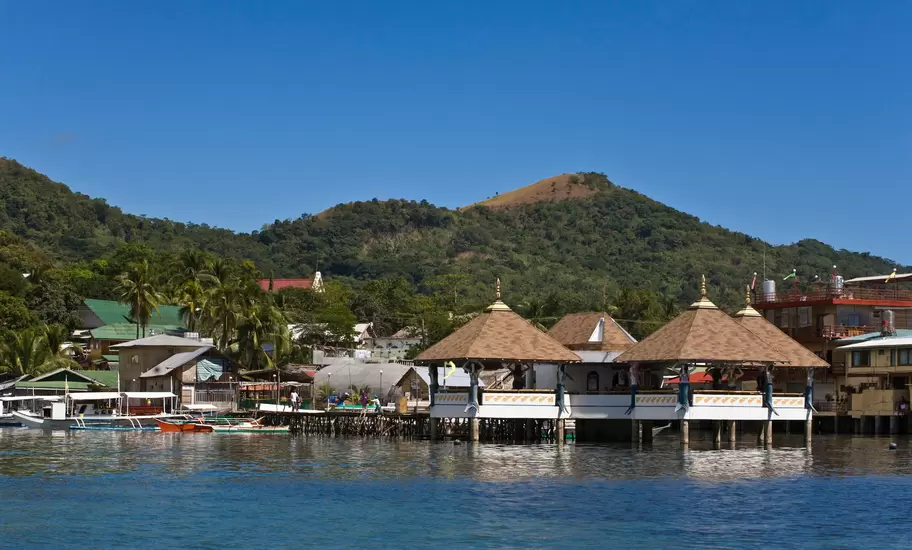 Stilted waterfront pavilions with pyramid roofs and outrigger boats below green hills, Coron Town, Palawan, Philippines