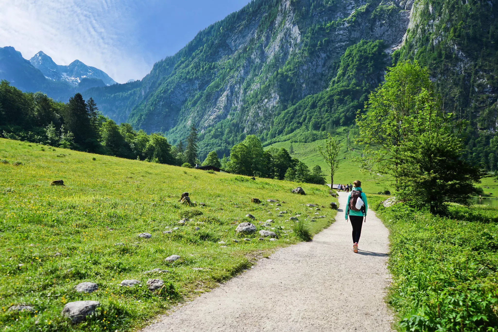 A solo hiker is pictured walking past fields on a gravel trail. The rocky slopes and peaks of mountains are seen in the distance.