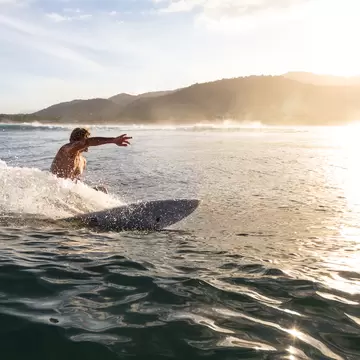 Surfer leaning into a turn on a wave, spray flying, backlit by the low sun with green hills on the horizon.