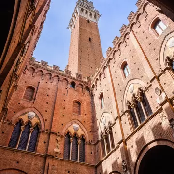 Looking up from a shaded arcade at the Torre del Mangia in Siena, framed by crenellated brick walls with Gothic windows.