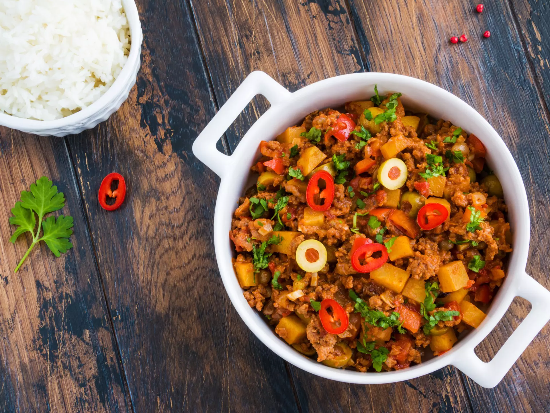 Classic Cuban dish picadillo a la habanera, a soft, fragrant stew of ground beef and tomatoes, with raisins and olives, in a white casserole and boiled rice on the wooden rustic table, top view.