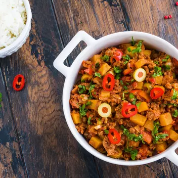 Classic Cuban dish picadillo a la habanera, a soft, fragrant stew of ground beef and tomatoes, with raisins and olives, in a white casserole and boiled rice on the wooden rustic table, top view.