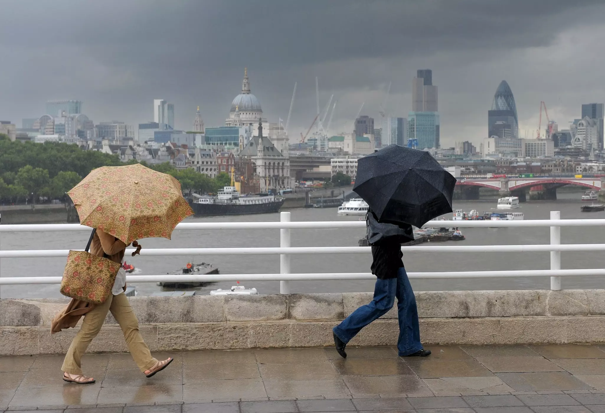 Two people shielded by umbrellas cross a bridge with a view of central London.