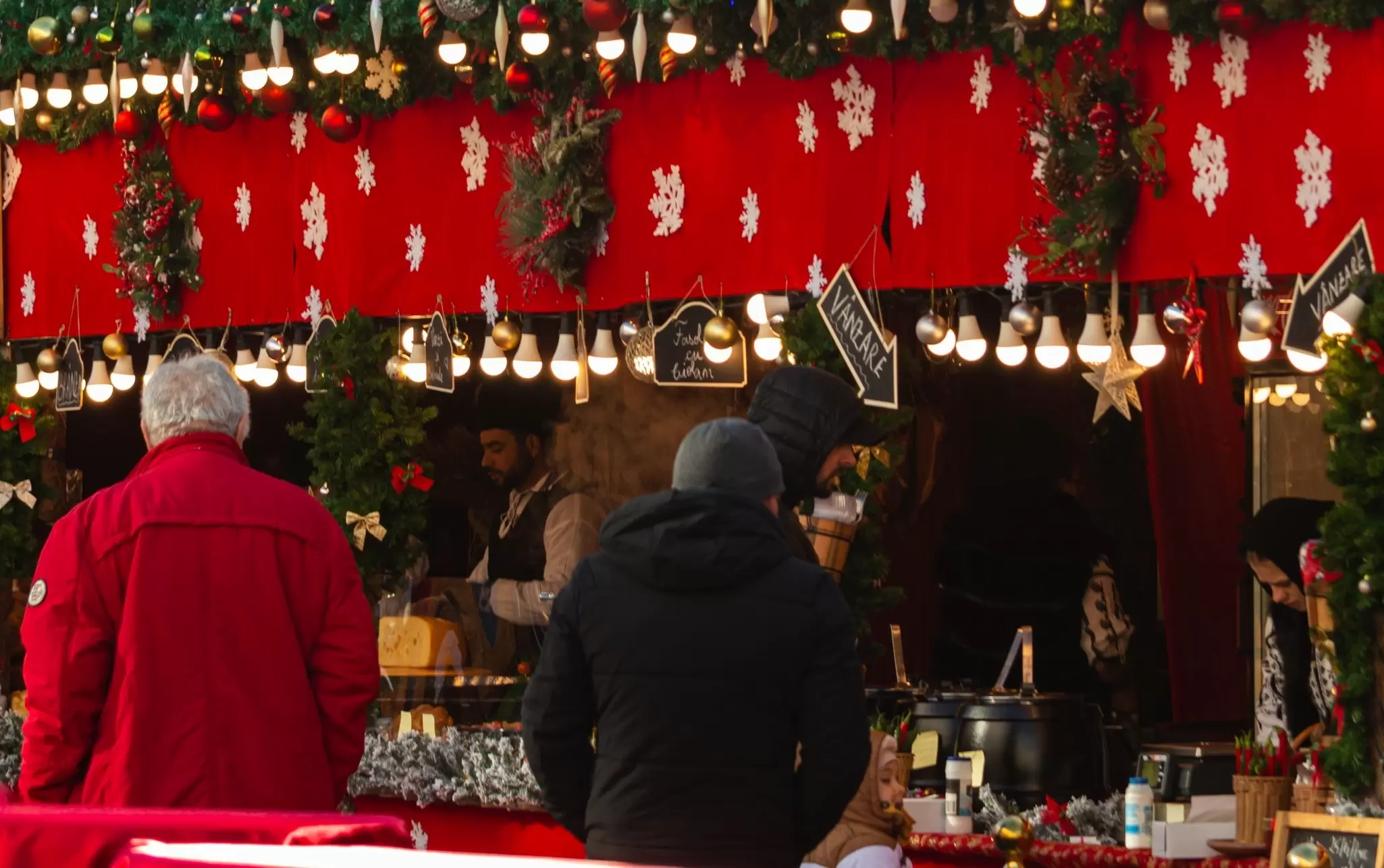 Bundled-up shoppers at a festive Christmas market food stall decorated with red cloth, white snowflakes, string lights and ornaments.