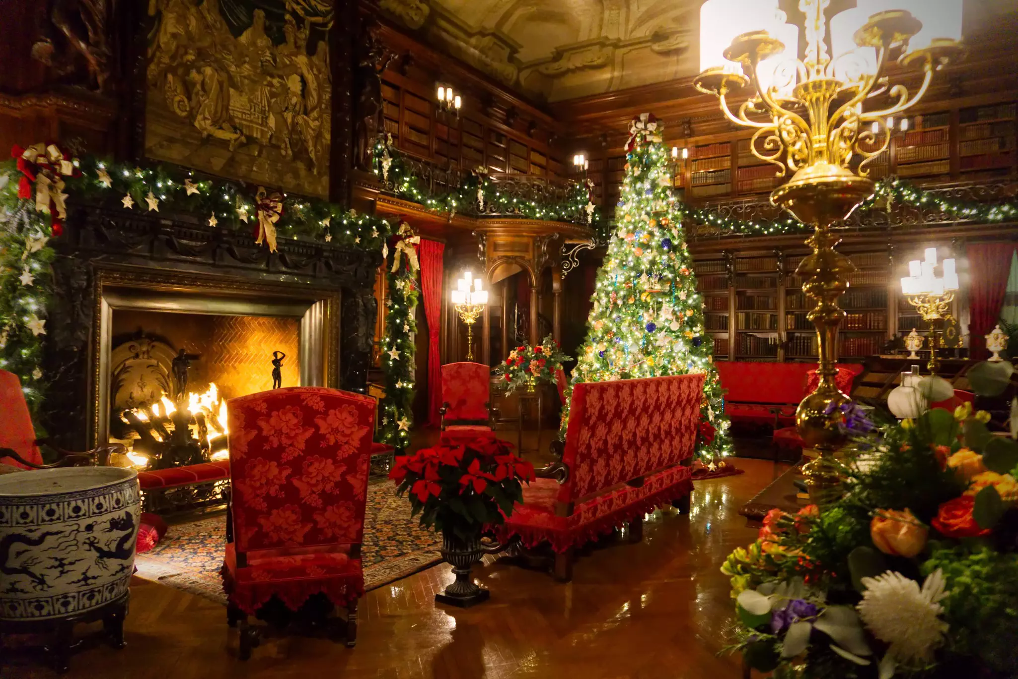 A room with shiny brown floors, seats upholstered in red and a large, lit fireplace is decorated for Christmas. A large, bright Christmas tree is in one corner