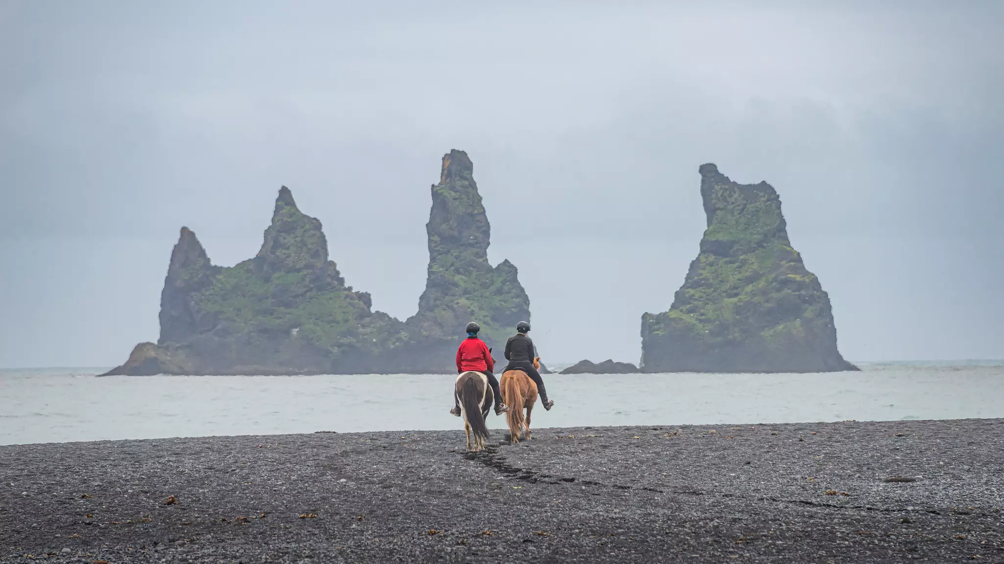 Two people ride horses on a black-sand beach. Basalt stacks rise dramatically from the sea in the distance.