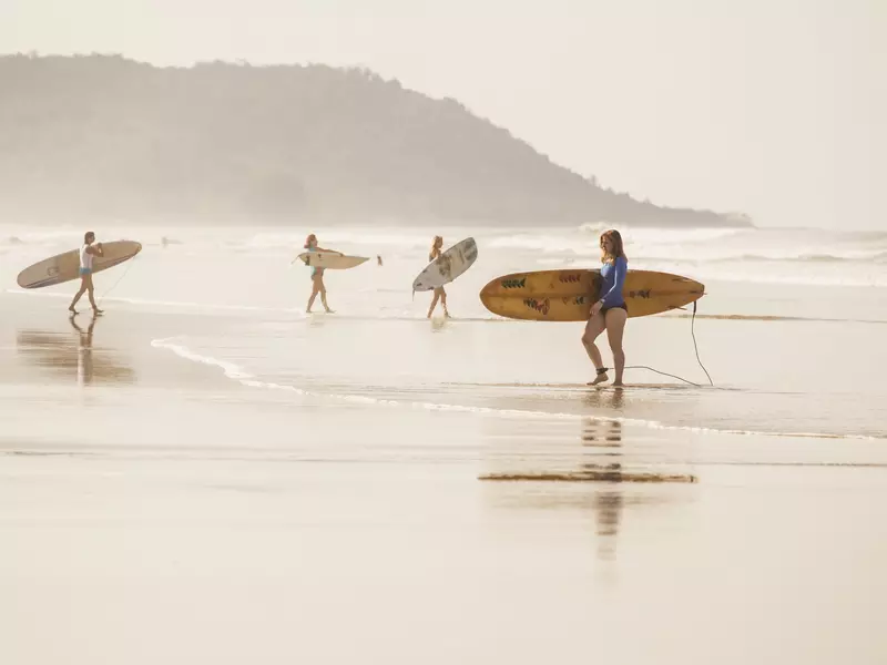 Four female surfers carry boards on the shore of a beach on a misty morning.