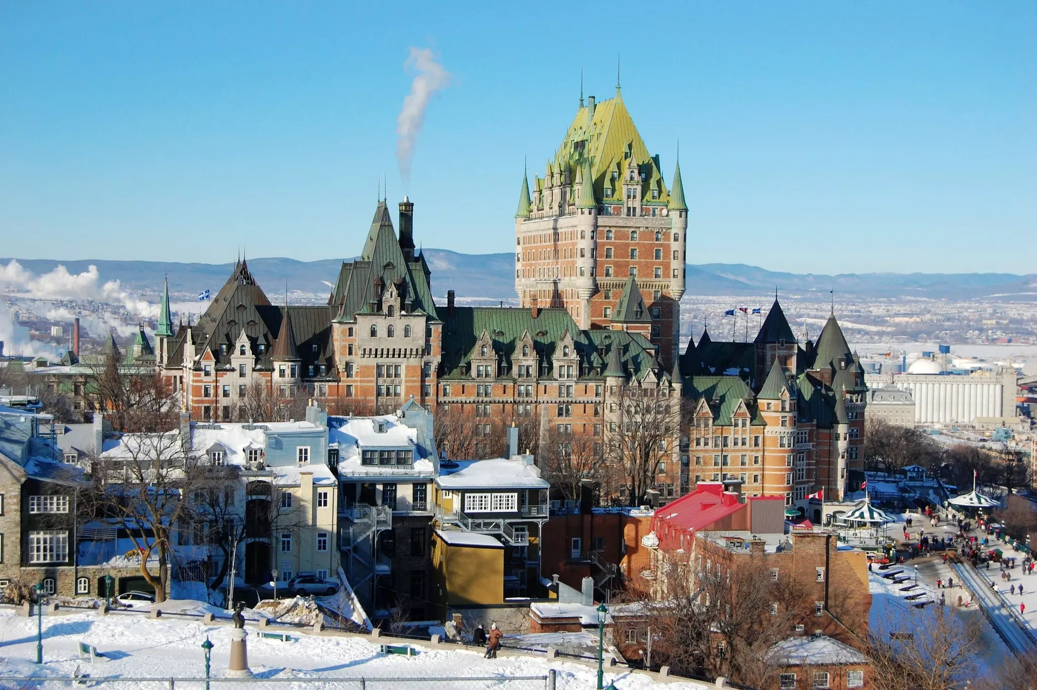 Wide shot of large brick castle with green roof with hills in the distance and snow and rowhouses in the foreground.