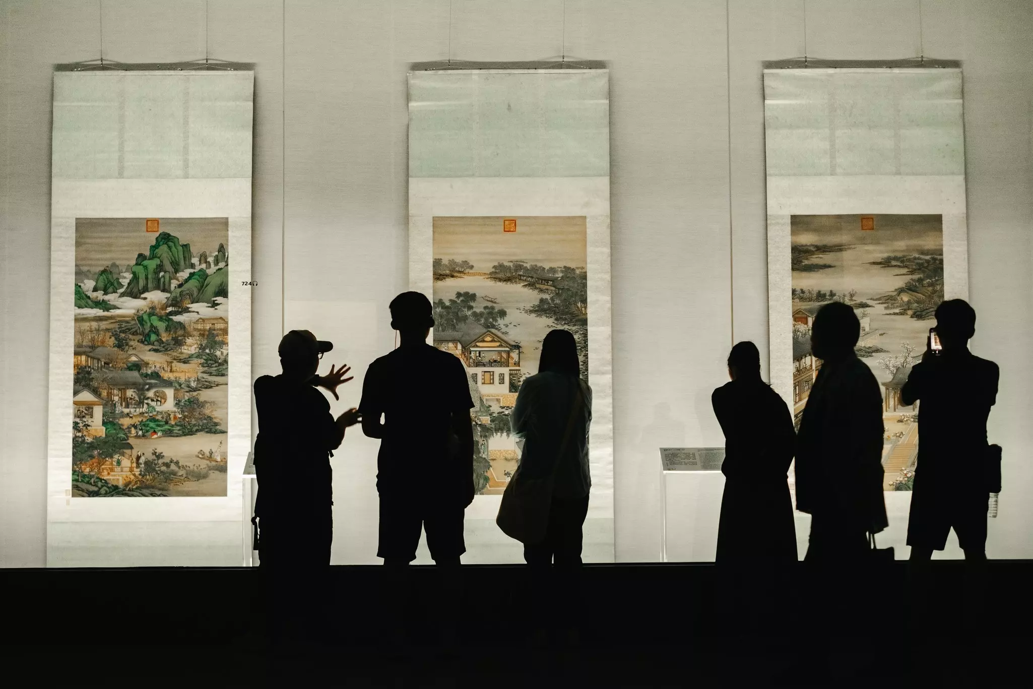 People in an art gallery are seen in silhouette. Painted scrolls hang from the walls.