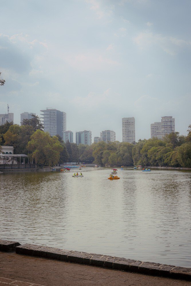 Pedal boats on a calm park lake lined with trees, with modern apartment towers behind under a hazy sky.