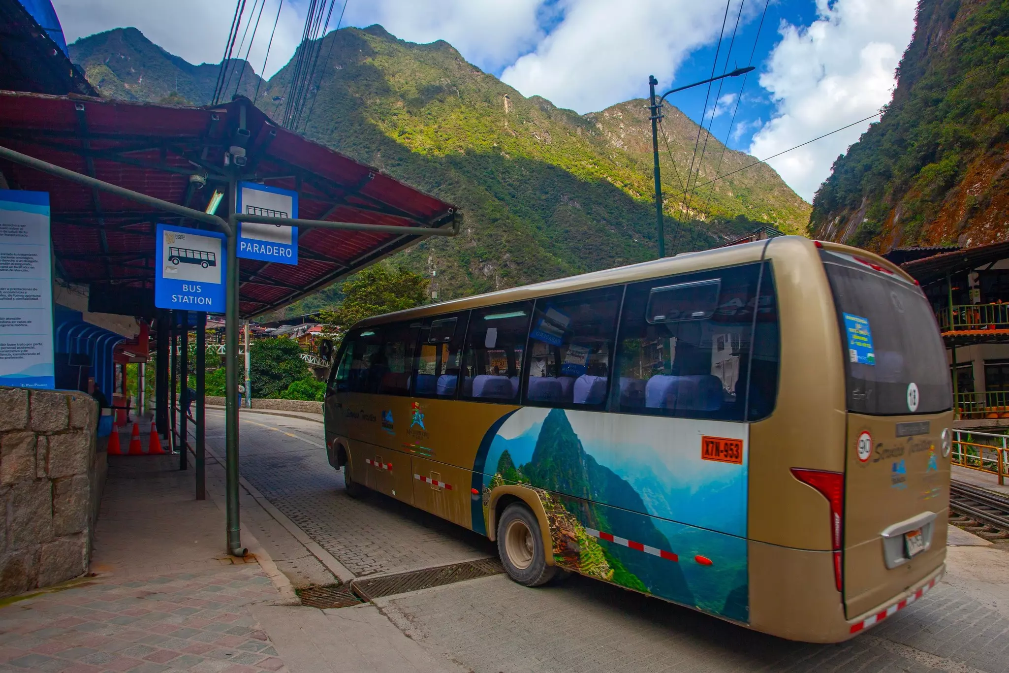 A coach is parked at a station in town. Steep green hillsides are seen in the immediate distance.