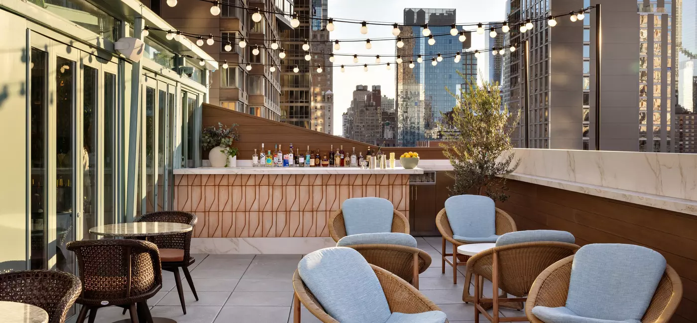 Seats with cushions are arranged around tables on a roofdeck under strings of lights in the daytime; tall buildings are in the background.