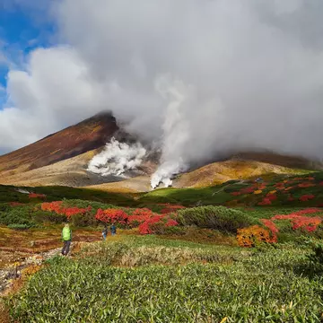 A hiker on a path in a meadow leading to a mountain shrouded in steam from vents in the earth.