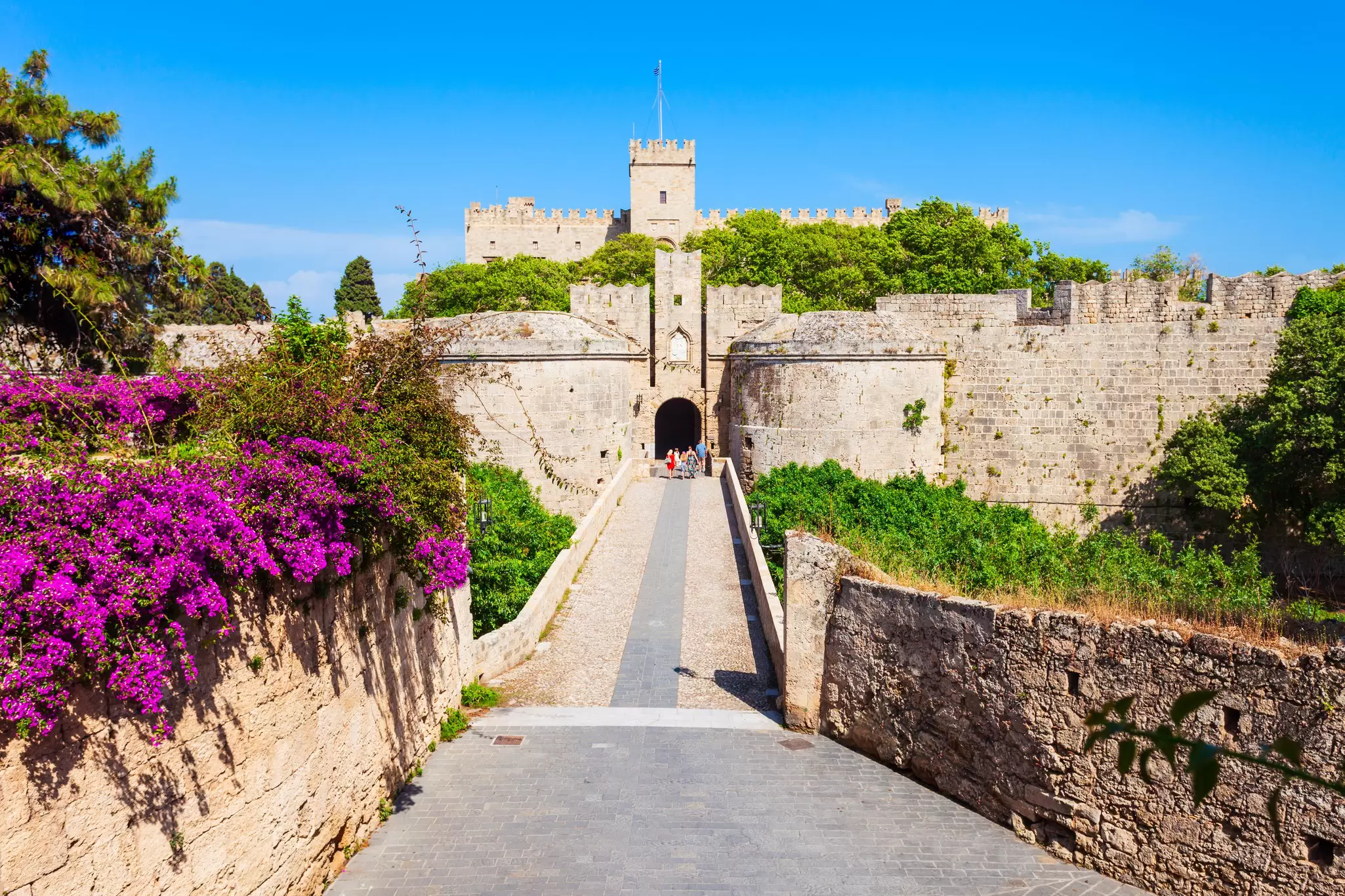 People walking towards a small arched entrance through a large stone, castle wall with a tower behind it.
