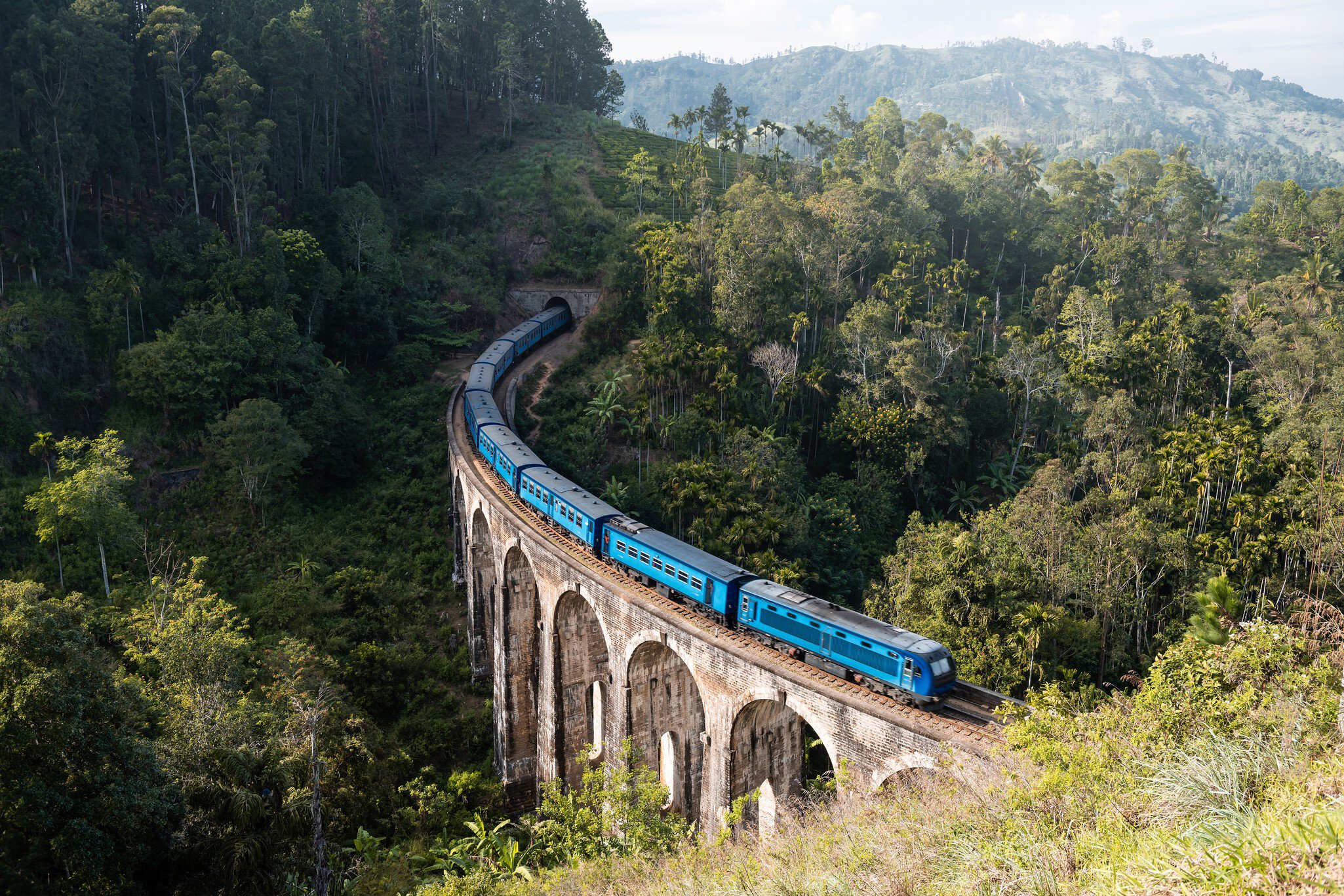 A blue train crosses a large stone bridge with nine arches over dense jungle.