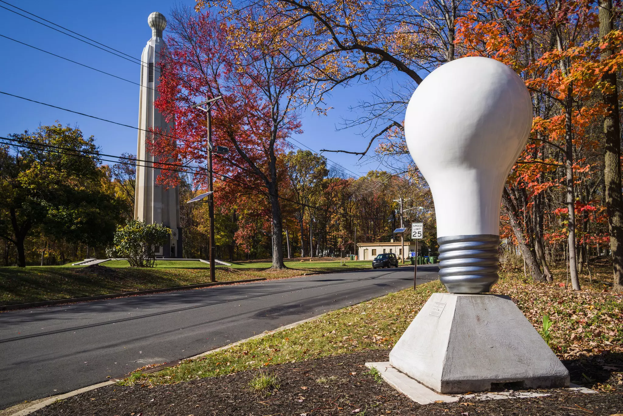 A large model of a lightbulb in the foreground and a monument tower across the road in the background with fall foliage in trees between them