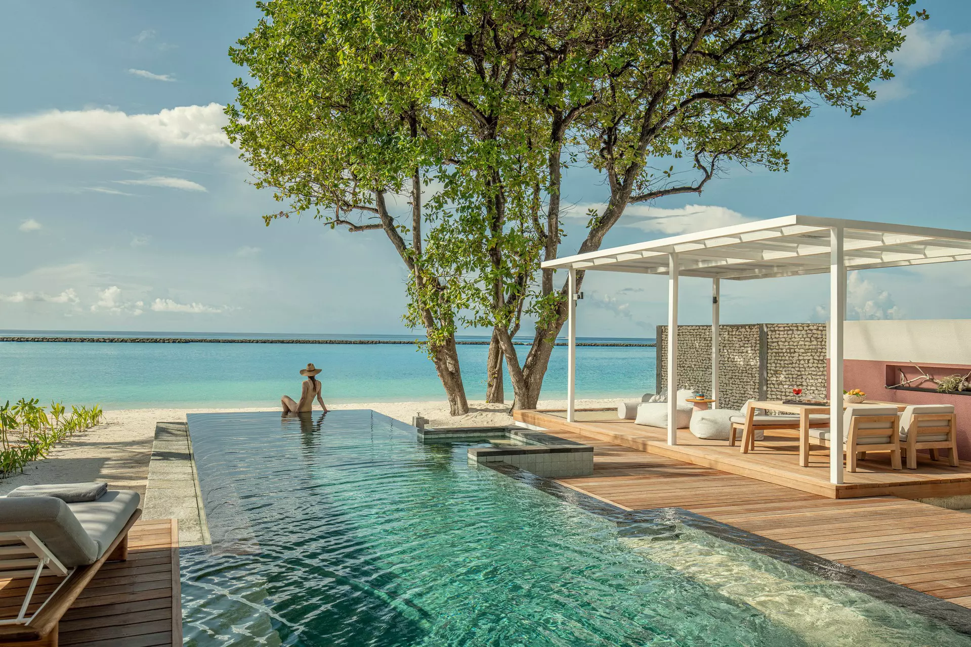 A woman in a sun hat sits at the edge of an infinity pool looking out at the ocean