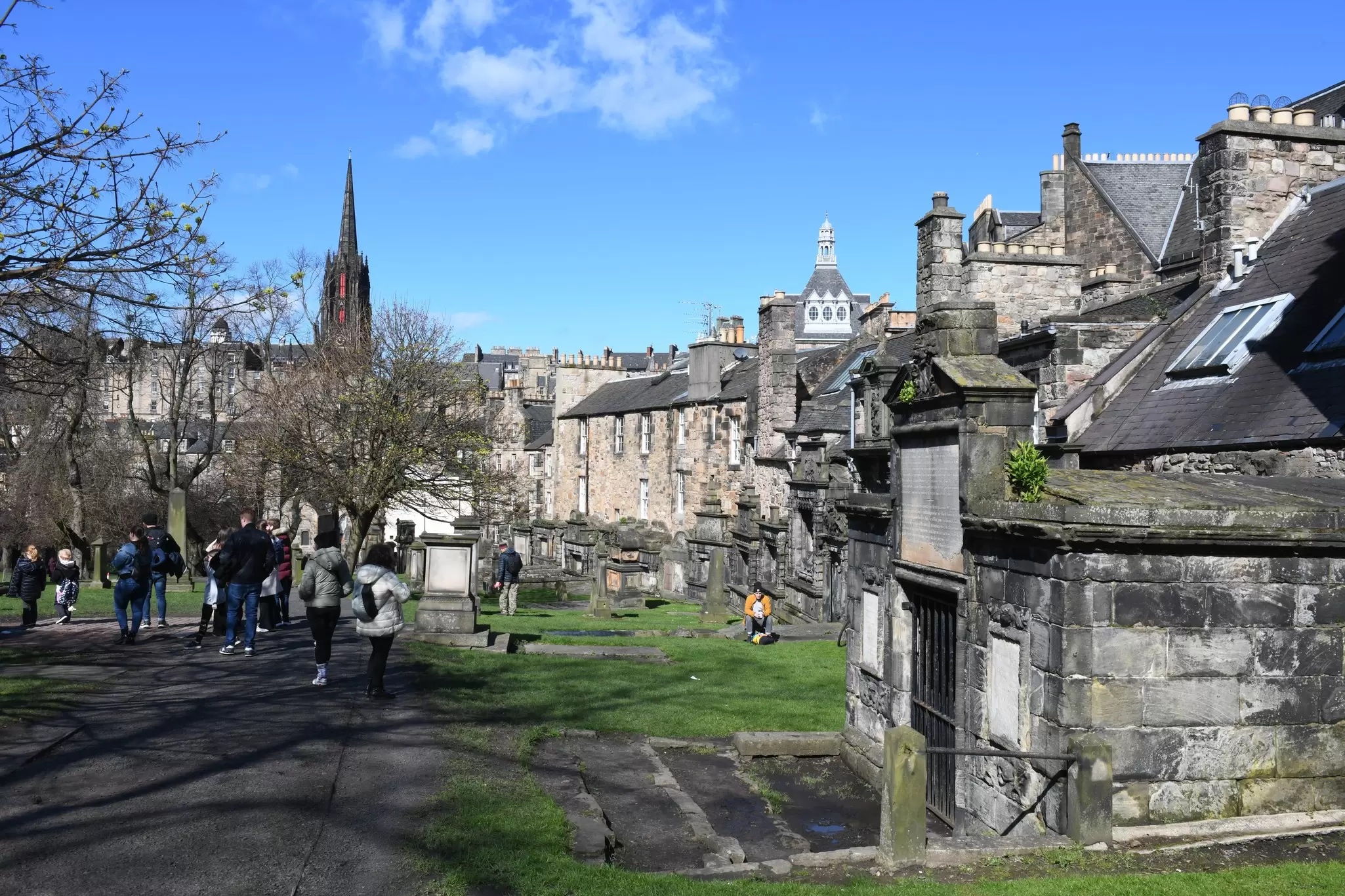 Visitors stroll through Greyfriars Kirkyard cemetery in Edinburgh on a sunny day.