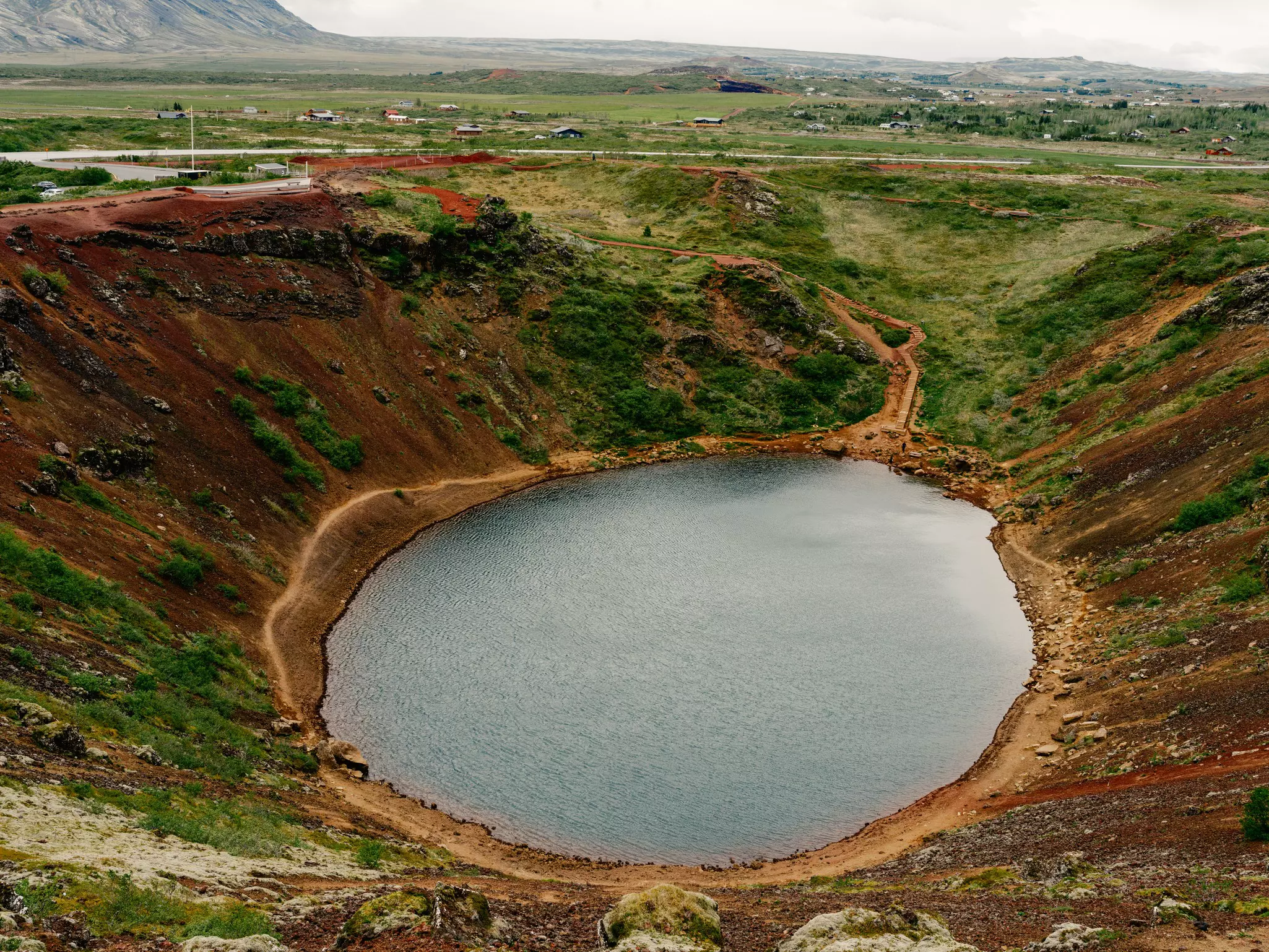 Large water-filled crater surrounded by grass- and dirt-covered hillside on an overcast day.