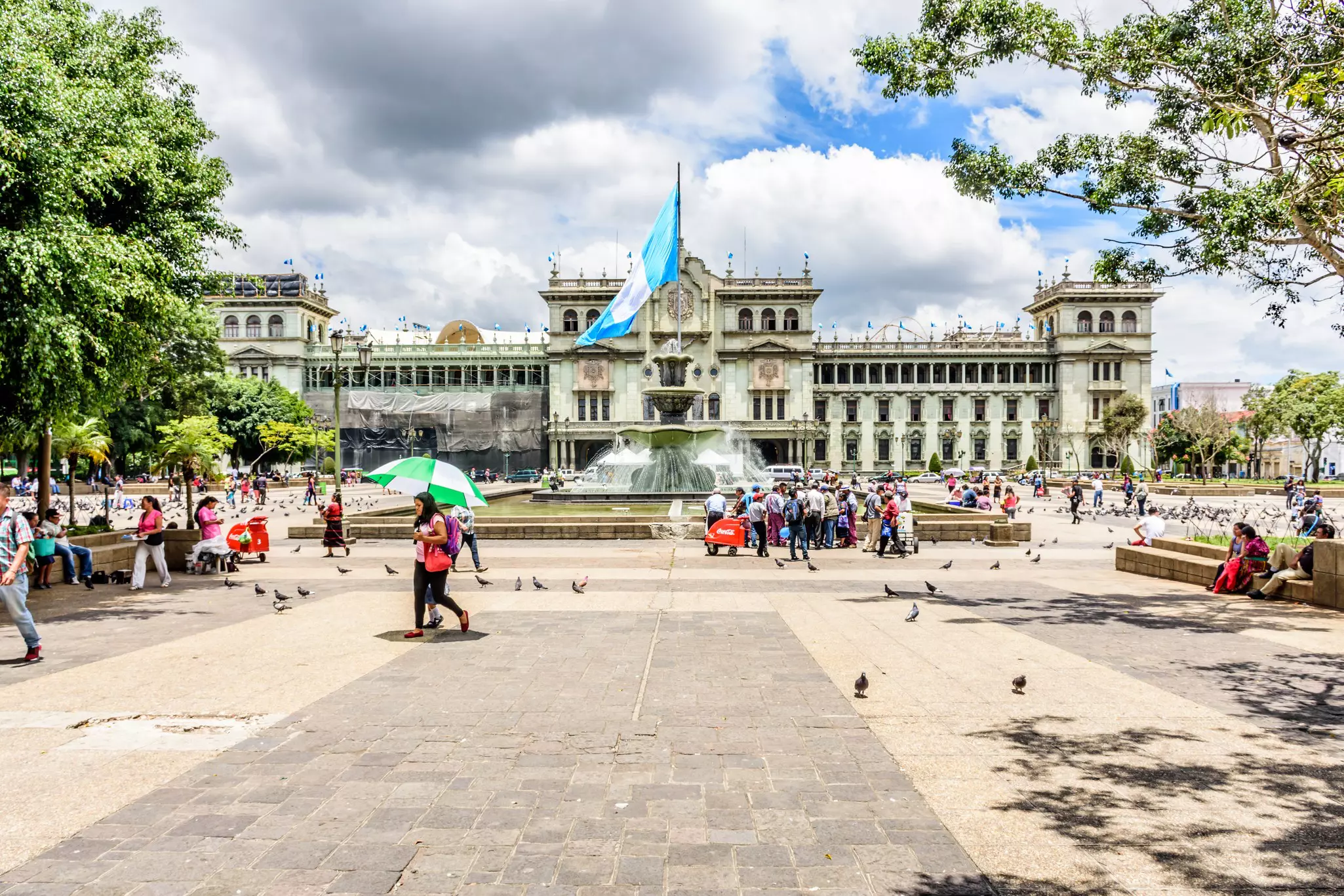 A busy city square with people passing by a central fountain and a flag pole flying the blue and white flag of Guatemala.
