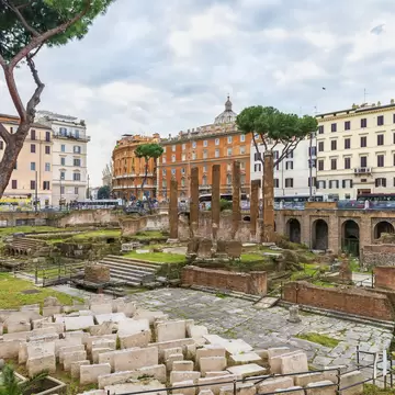 Sunken excavation of column stumps and stone blocks below street level, ringed by pastel apartment blocks and pines.