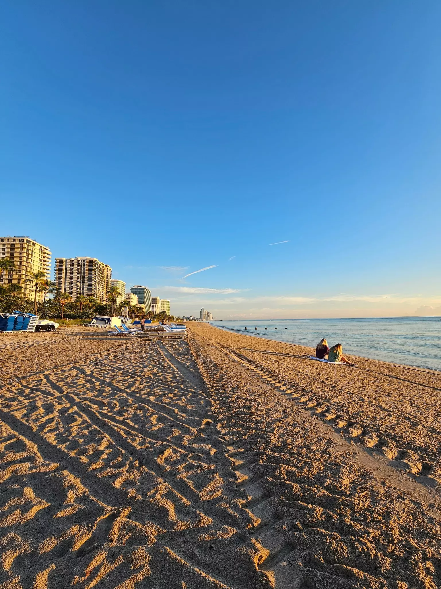 a stunning summer landscape along the beach at sunrise with blue and white lounge chairs, blue ocean water, palm trees, hotels, and footprints in the sand at Bal Harbour Beach in Miami Beach Florida