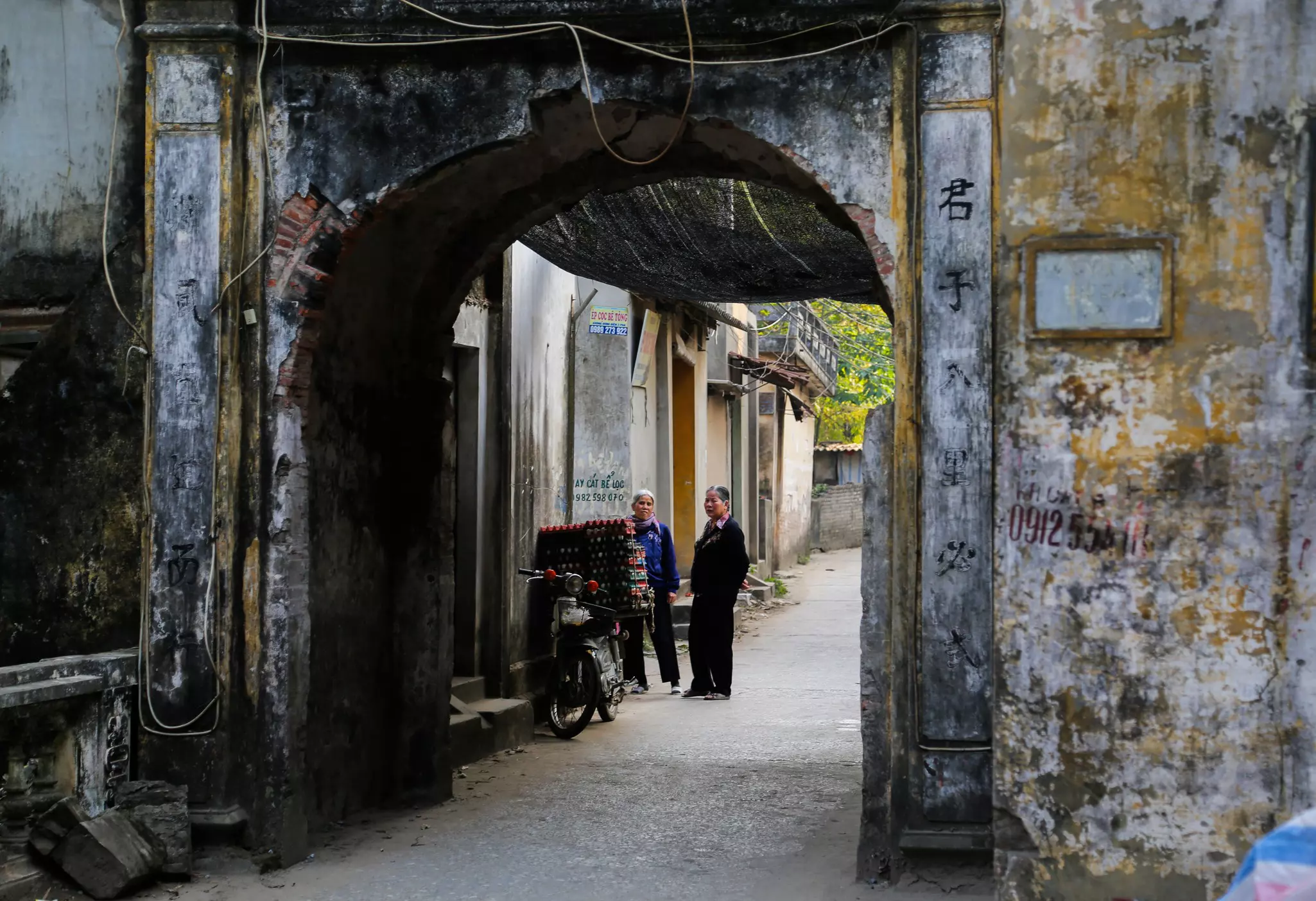 Two people standing next to a motorbike near a weathered arch with lettering and numbers etched into it