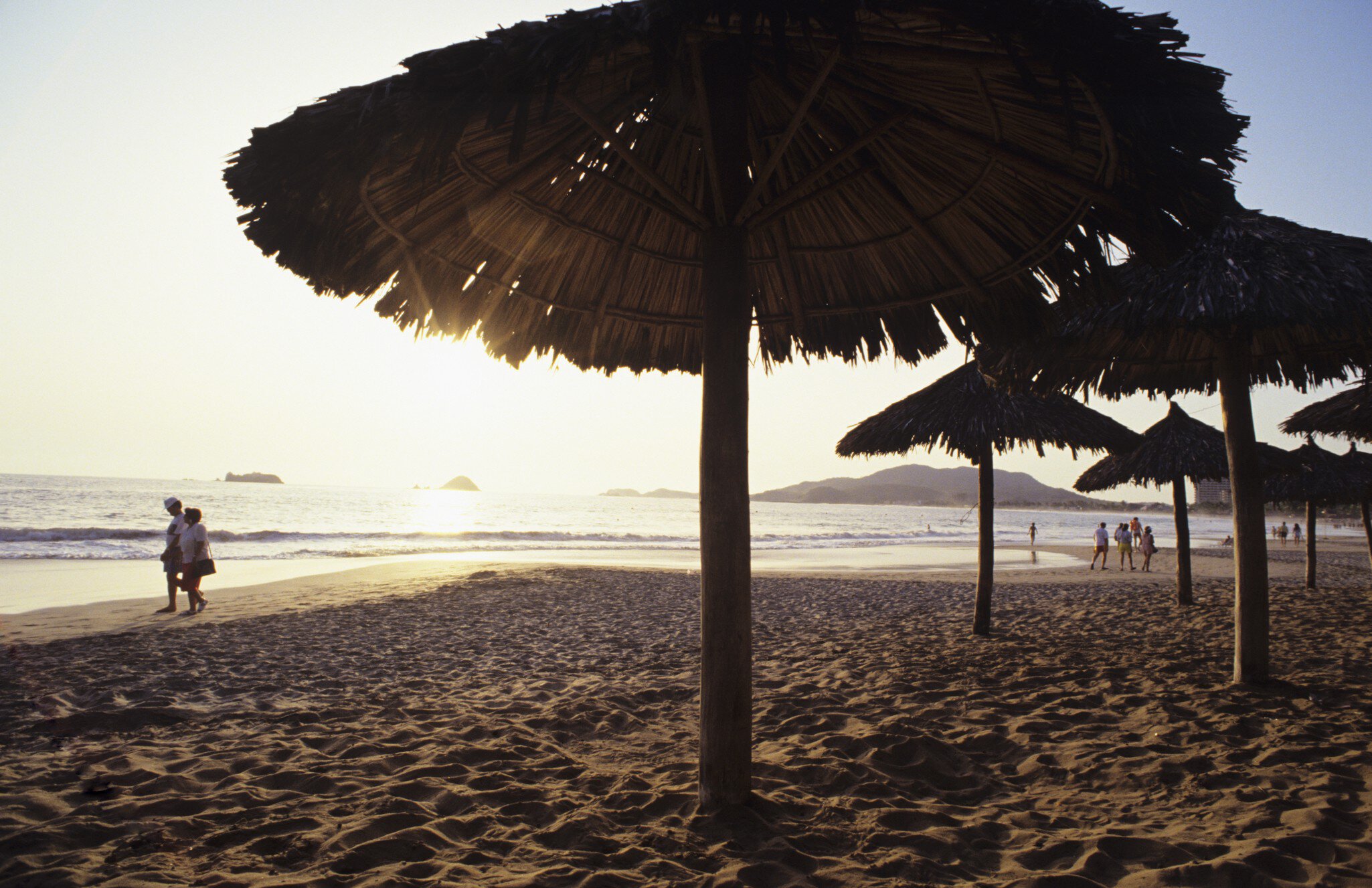 Mexico, Ixtapa, ocean sunset with couples walking along shoreline, umbrellas in sand.