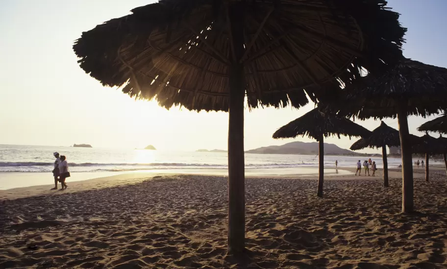 Thatched palapa umbrellas on a sandy beach silhouetted against a hazy sunset over the sea, with people strolling by the water.