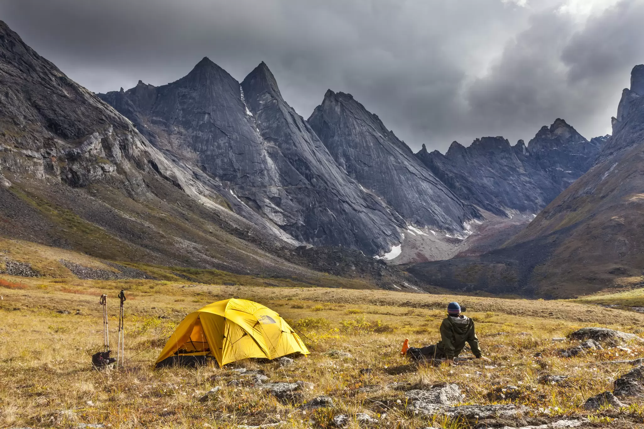 A person sits on the ground next to a yellow tent looking at a rocky mountain range.