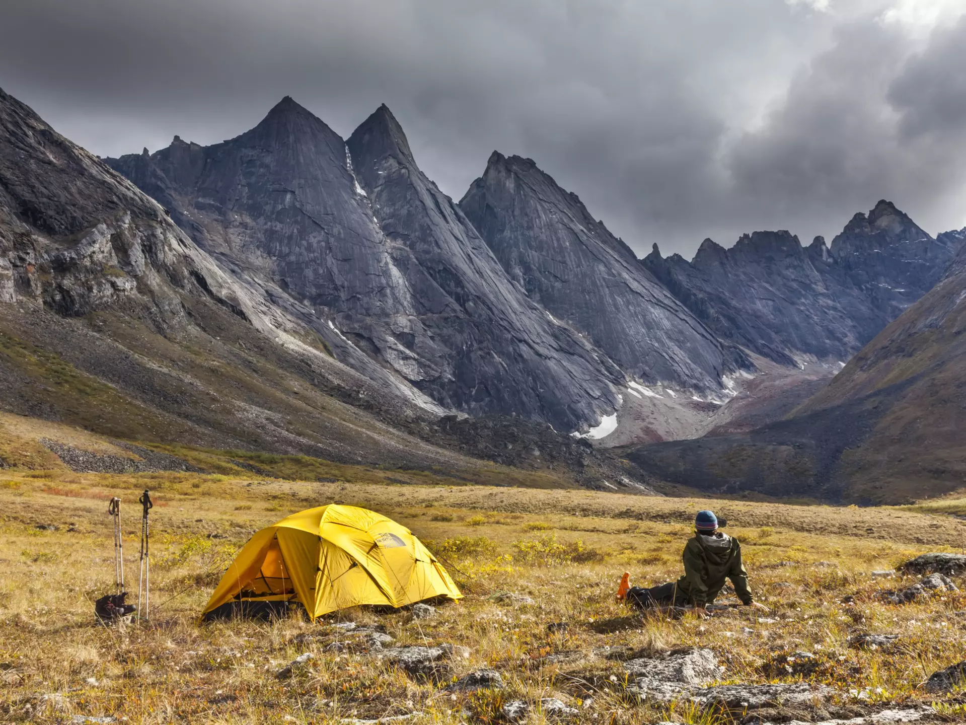 A person sits on the ground next to a yellow tent looking at a rocky mountain range.
