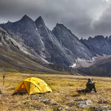 A person sits on the ground next to a yellow tent looking at a rocky mountain range.