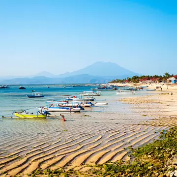 Rippled sand and seaweed at low tide with moored outrigger boats, a person wading and a volcano on the horizon.