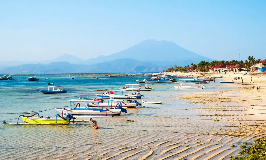 Rippled sand and seaweed at low tide with moored outrigger boats, a person wading and a volcano on the horizon.
