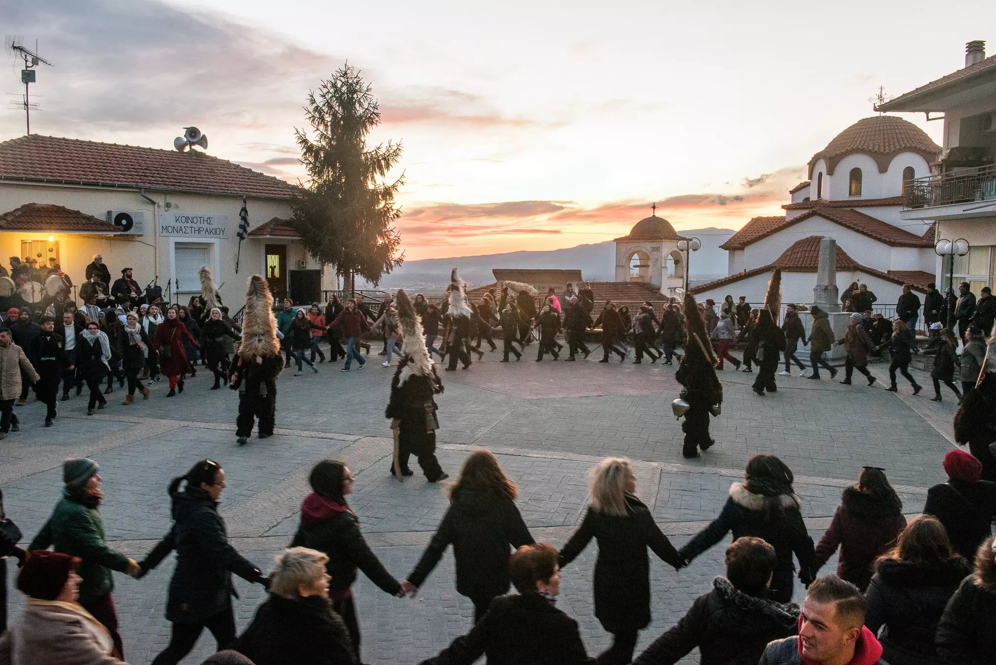Dancers join hands and circle around an open plaza at sunset. Several costumed figures are in the middle of the circle.