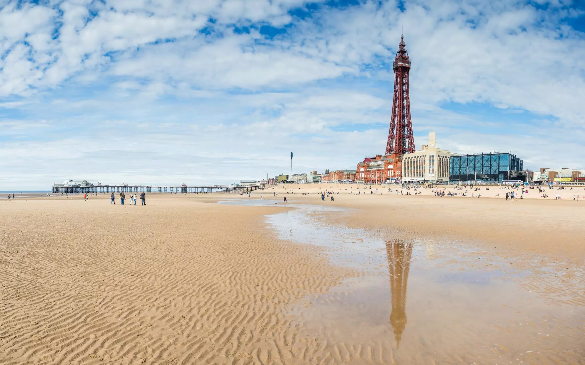 A distinctive tower rises above buildings that line the shorefront near a sandy beach. A long pier is in the distance.