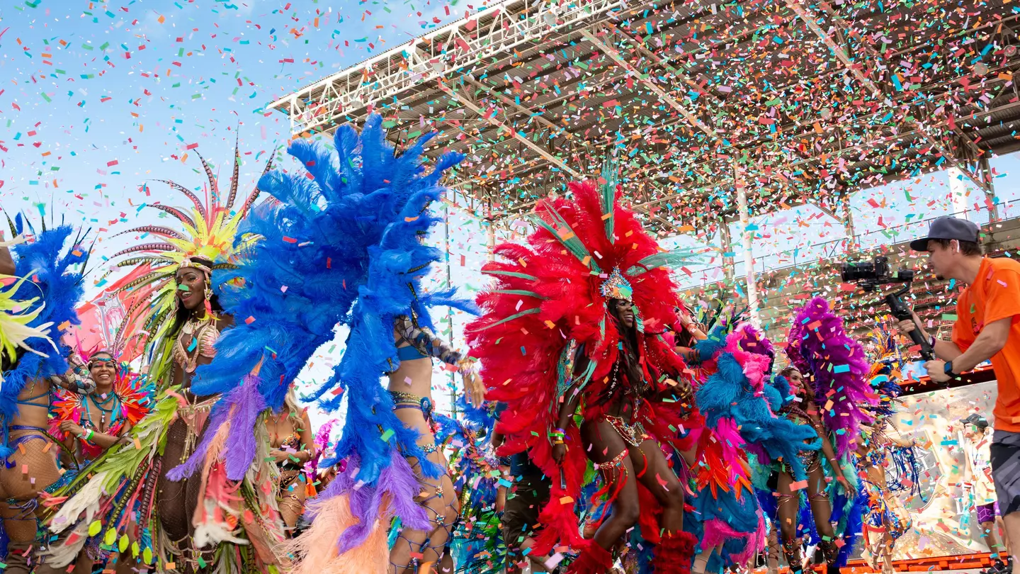 Masqueraders in feathered costumes dance in a stadium as confetti falls around them.