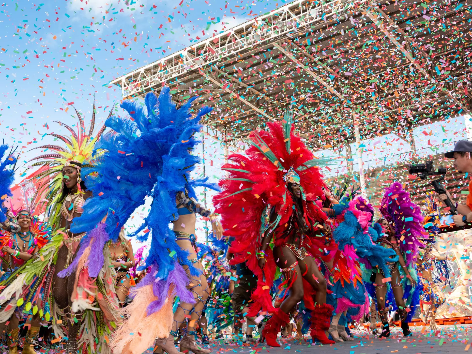 Masqueraders in feathered costumes dance in a stadium as confetti falls around them.