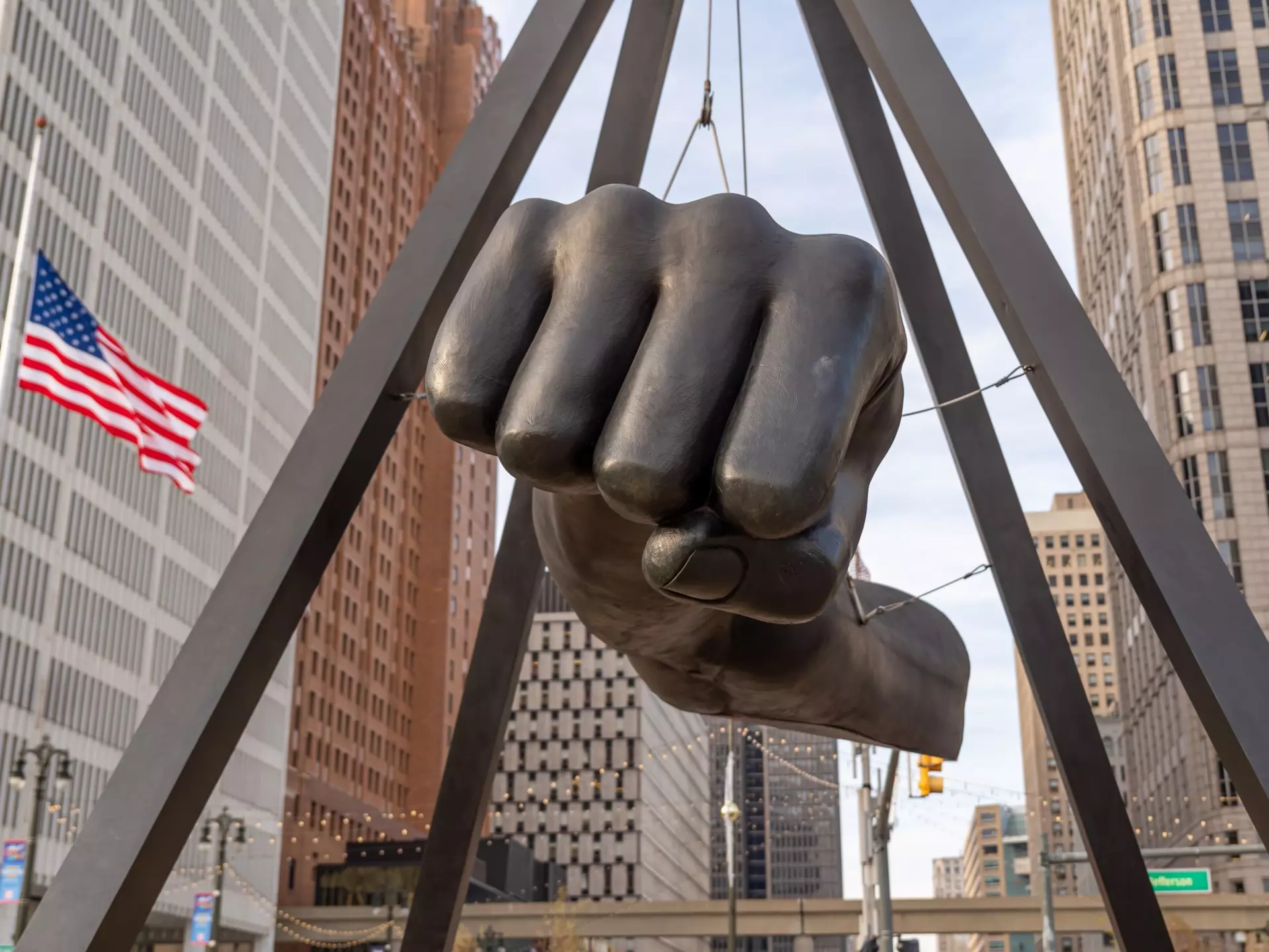 A large bronze fist sculpture suspended between bronze columns with city buildings and an American flag in the distance.