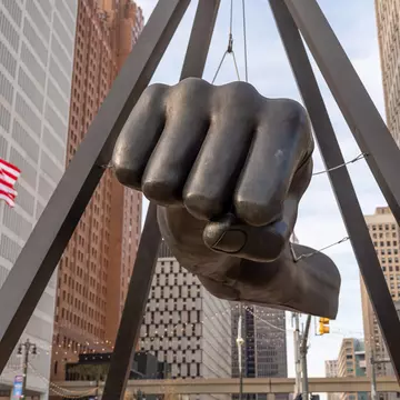 A large bronze fist sculpture suspended between bronze columns with city buildings and an American flag in the distance.